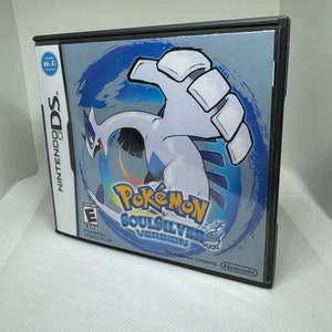 Puede incluir: Una caja de juego de Nintendo DS negra con una imagen azul y blanca de un personaje Pokémon en la parte delantera. El juego se llama "Pokémon Versión SoulSilver" y presenta el texto "The Pokémon Company" y "Nintendo".