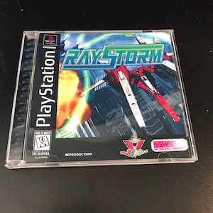 以下が含まれることがあります： レイストームのPlayStationゲームケース。赤い白の宇宙船がオレンジと緑の爆発を背景に都市を飛行している。ケースには「レイストーム」と表示され、ESRBから「Kids to Adults」の評価「KA」を受けている。