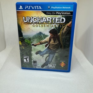 Puede incluir: Una caja de juego PlayStation Vita azul con una imagen de un hombre colgado de una liana. El juego es Uncharted: Golden Abyss. El juego está clasificado como T para adolescentes por la ESRB.