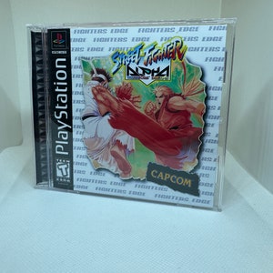 Może przedstawiać: Pudełko gry PlayStation do gry Street Fighter Alpha: Warriors' Dreams. Pudełko jest białe z czerwonym, niebieskim i zielonym obrazkiem dwóch walczących postaci. Na pudełku widnieje tekst "Street Fighter Alpha" i "Warriors' Dreams". Pudełko zawiera również logo Capcom i oznaczenie dla młodzieży.
