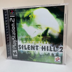 May include: A PlayStation 2 video game case with a green and black design. The front of the case features a woman's face with a blurry background. The title of the game is "Silent Hill 2" in white text with a red and black logo. The ESRB rating is "M" for Mature 17+.