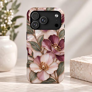 Puede incluir: Una funda para smartphone con un patrón floral estilo vidriera, con pétalos de color rosa claro y ciruela oscuro, hojas verde salvia y contornos dorados. El diseño incluye un módulo de cámara negro en la parte superior con tres lentes.