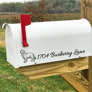 May include: A white metal mailbox featuring a bright red flag on the left side. The side of the mailbox displays a black line drawing of a dog standing in profile, followed by the black text 1704 Burberry Lane in a cursive script font.