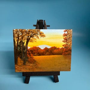 May include: Miniature landscape painting on a small wooden easel. The artwork depicts a fall scene with trees in shades of orange and yellow, a field, and a sunset. The easel is black.