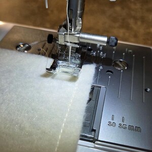 K&ouml;nnte beinhalten: Nahaufnahme einer N&auml;hmaschine in Betrieb, die ein St&uuml;ck wei&szlig;en Stoffes n&auml;ht. Die Nadel befindet sich &uuml;ber dem Stoff, der Faden ist sichtbar. Die Metallplatte der Maschine hat Ma&szlig;e in mm.