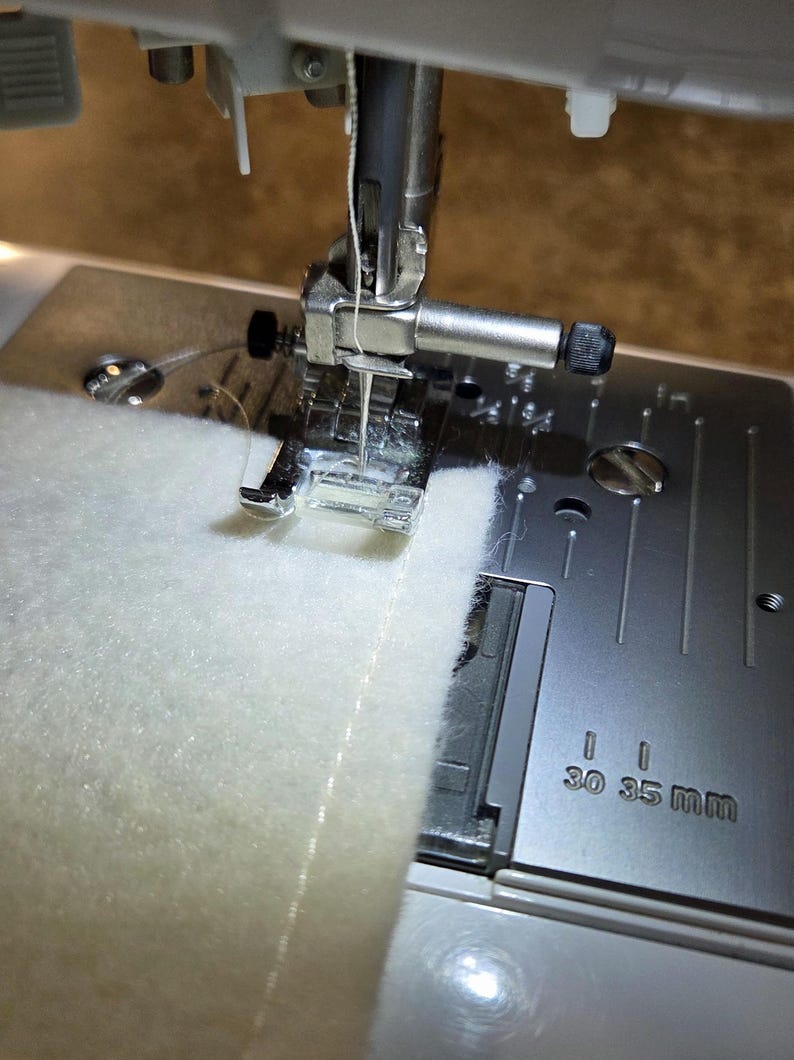 K&ouml;nnte beinhalten: Nahaufnahme einer N&auml;hmaschine in Betrieb, die ein St&uuml;ck wei&szlig;en Stoffes n&auml;ht. Die Nadel befindet sich &uuml;ber dem Stoff, der Faden ist sichtbar. Die Metallplatte der Maschine hat Ma&szlig;e in mm.