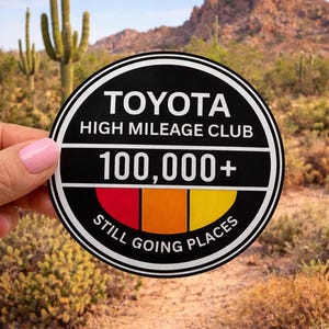 May include: A black and white Toyota High Mileage Club sticker with the text "100,000+" and "Still Going Places." The sticker has red, orange, and yellow color bars. The background shows a desert landscape with cacti and mountains.