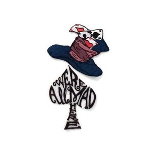 Op de afbeelding: Een zwarte schoppen speelkaart met de woorden "We're All Mad Here" geschreven in de vorm. Een blauwe en rode hoed met speelkaarten erop bevindt zich boven de schoppen.