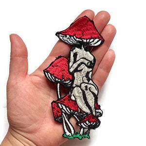 May include: An embroidered patch featuring a nude figure sitting on three red mushrooms with white spots. The mushrooms have green stems and leaves.