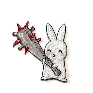 Pode incluir: Patch bordado com um coelho branco sorridente segurando um porrete cinza com espinhos vermelhos. O coelho tem bochechas rosadas e olhos pretos. O patch está em um fundo branco.
