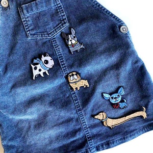 Op de afbeelding: Een blauwe denim overalljurk met vijf geborduurde hondenvlekken. De vlekken zijn van een Franse bulldog, een mops, een teckel, een bulldog en een chihuahua.