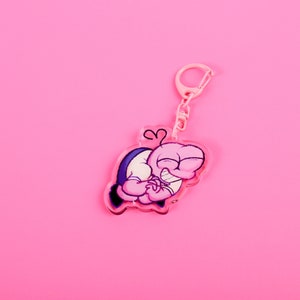 Puede incluir: Un llavero rosa con un dise&ntilde;o de personaje de dibujos animados. El personaje es rosa con un atuendo azul y blanco, y se abraza a s&iacute; mismo. El llavero tiene un cierre y una cadena rosas, sobre un fondo rosa.