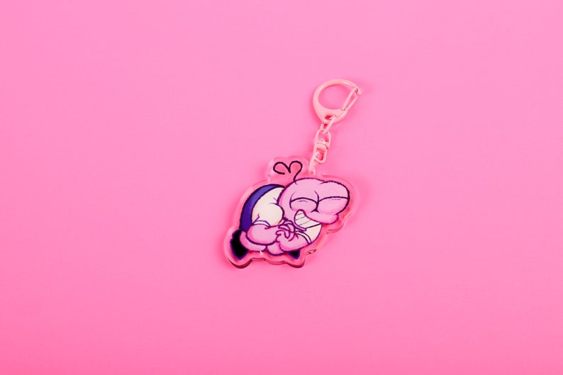Puede incluir: Un llavero rosa con un dise&ntilde;o de personaje de dibujos animados. El personaje es rosa con un atuendo azul y blanco, y se abraza a s&iacute; mismo. El llavero tiene un cierre y una cadena rosas, sobre un fondo rosa.