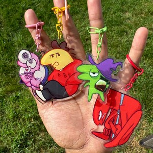Puede incluir: Una mano muestra cinco llaveros de personajes de dibujos animados con anillos de colores. Los llaveros presentan varios personajes en rojo, amarillo, verde, rosa y morado. El fondo es hierba verde.
