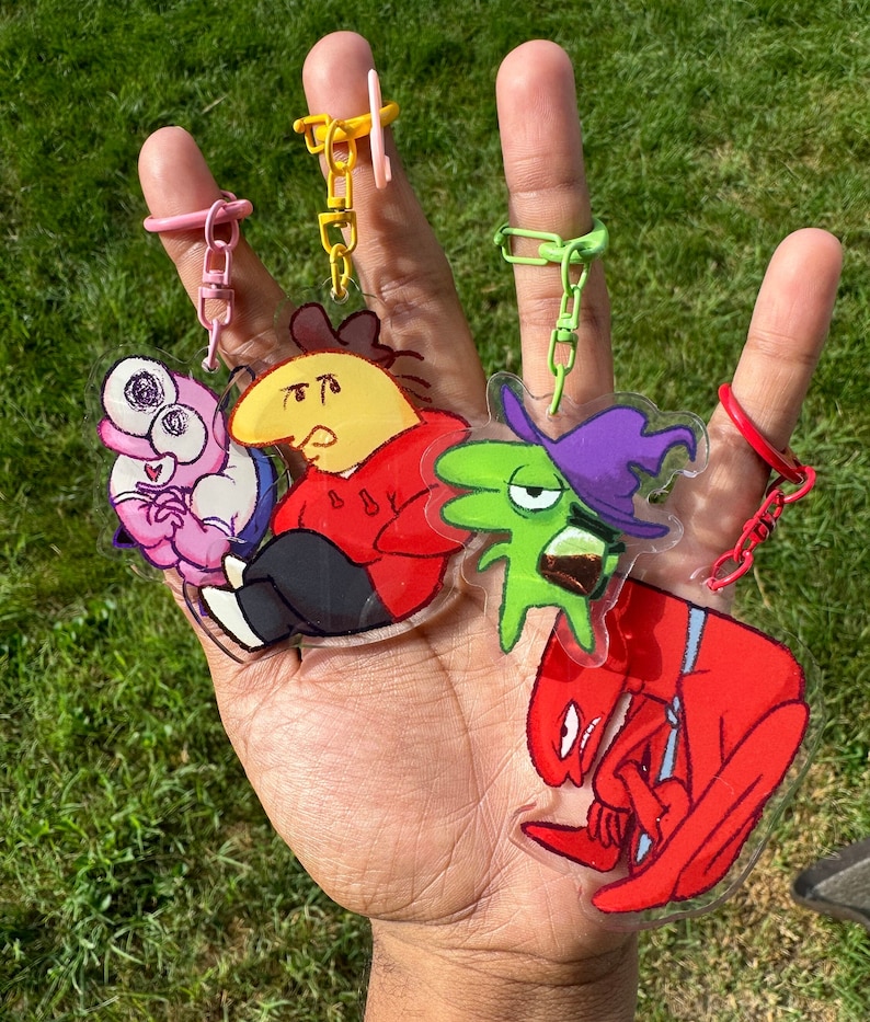 Puede incluir: Una mano muestra cinco llaveros de personajes de dibujos animados con anillos de colores. Los llaveros presentan varios personajes en rojo, amarillo, verde, rosa y morado. El fondo es hierba verde.