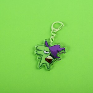 Puede incluir: Un llavero acr&iacute;lico verde con un personaje de bruja de dibujos animados con un sombrero morado y una bebida. El llavero tiene un cierre plateado y un fondo verde.