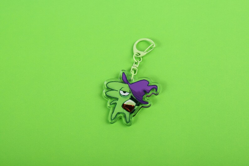 Puede incluir: Un llavero acr&iacute;lico verde con un personaje de bruja de dibujos animados con un sombrero morado y una bebida. El llavero tiene un cierre plateado y un fondo verde.