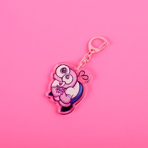 Puede incluir: Un llavero rosa con un personaje de dibujos animados con ojos grandes, una camisa blanca y detalles azules. El llavero tiene un cierre y una cadena rosas, sobre un fondo rosa brillante. El dise&ntilde;o es caprichoso y juguet&oacute;n.