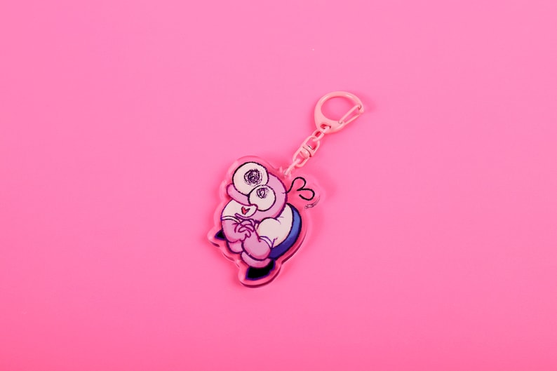 Puede incluir: Un llavero rosa con un personaje de dibujos animados con ojos grandes, una camisa blanca y detalles azules. El llavero tiene un cierre y una cadena rosas, sobre un fondo rosa brillante. El dise&ntilde;o es caprichoso y juguet&oacute;n.