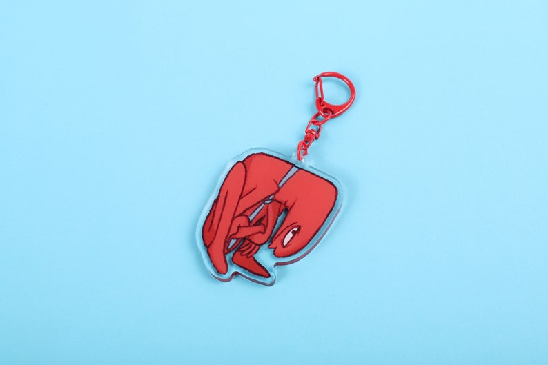 Puede incluir: Un llavero acr&iacute;lico rojo con una figura de dibujos animados en posici&oacute;n encogida. El llavero tiene un cierre rojo y un borde transparente. El fondo es de color azul claro liso.