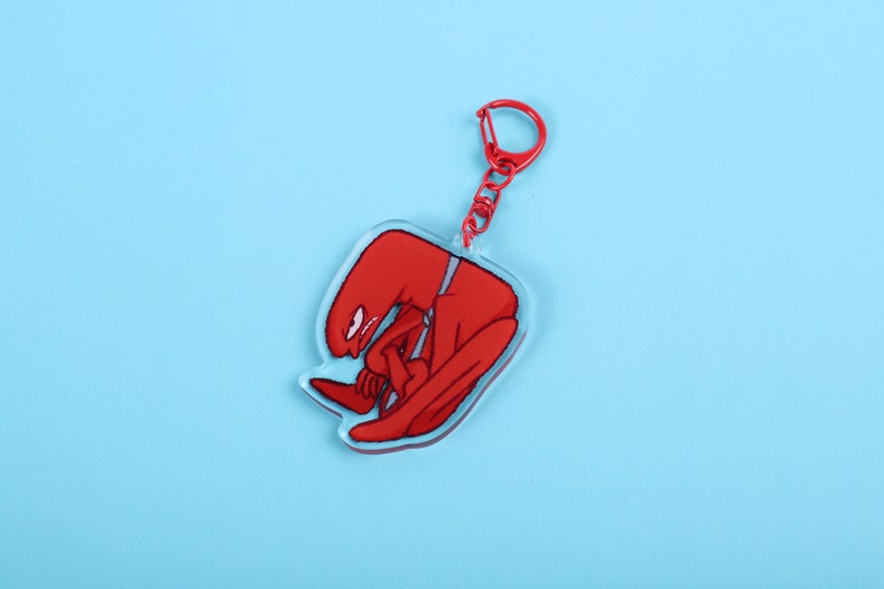 Puede incluir: Un llavero acr&iacute;lico rojo con una figura estilizada en posici&oacute;n sentada. La figura tiene un cuerpo largo y curvado y una cabeza peque&ntilde;a con un ojo blanco. El llavero tiene un cierre y una cadena de metal rojo, sobre un fondo azul claro.