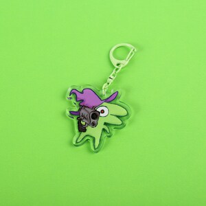 Puede incluir: Un llavero de acr&iacute;lico transparente con un personaje de dibujos animados verde que lleva un sombrero de bruja morado y sostiene un arma. El llavero tiene una cadena y un cierre de color plateado. El fondo es verde lima.