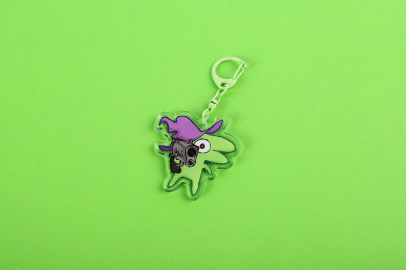 Puede incluir: Un llavero de acr&iacute;lico transparente con un personaje de dibujos animados verde que lleva un sombrero de bruja morado y sostiene un arma. El llavero tiene una cadena y un cierre de color plateado. El fondo es verde lima.