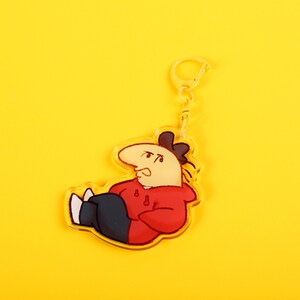 Puede incluir: Un llavero acr&iacute;lico transparente con un personaje de dibujos animados con una sudadera roja y pantalones azul oscuro. El personaje tiene una nariz grande y est&aacute; tumbado. El llavero tiene un cierre y un anillo dorados, sobre un fondo amarillo.