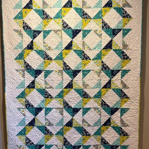 May include: A handmade quilt with a geometric pattern. The quilt features a white background with repeating diamond shapes made of teal, yellow, and navy blue triangles. The quilt is machine quilted with a floral pattern.