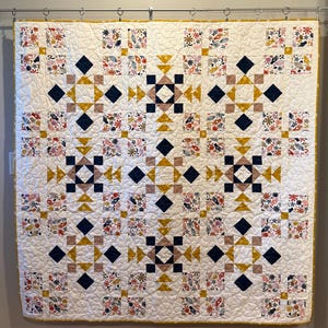 May include: A square quilt featuring a repeating geometric pattern of navy blue, mustard yellow, and soft pink squares and triangles on a white background. The fabric includes printed motifs of hands, flowers, and stars, with overall swirling quilting.