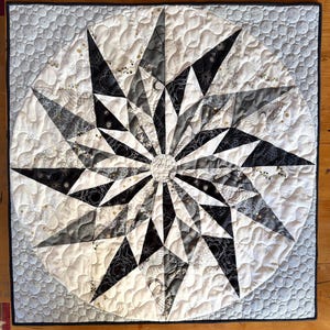 May include: A handmade quilt featuring a starburst design in black, white, and gray. The quilt has a circular pattern with pointed star shapes radiating from the center. The background is white with a quilted texture, and the edges are finished with a black border.