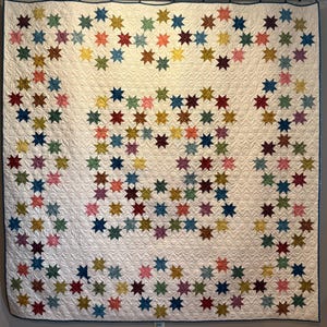 May include: A handmade quilt with a white background and a pattern of colorful star shapes. The stars are in shades of red, blue, green, yellow, and pink. The quilt is displayed on a wall.