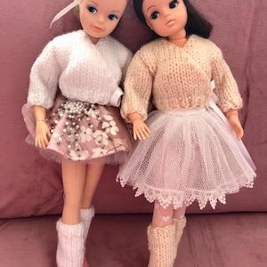 May include: Two dolls dressed in ballerina outfits. The doll on the left is wearing a white sweater, a pink skirt with floral print and white leg warmers. The doll on the right is wearing a beige sweater, a white tulle skirt with lace trim and beige leg warmers.