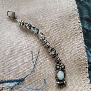 May include: A decorative owl-themed charm with an antique brass finish. The charm features iridescent teardrop-shaped embellishments and a pearlescent oval stone. It includes a lobster clasp for easy attachment.