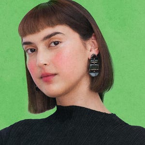 May include: A person wearing black geometric earrings with gold detailing. The person is wearing a black top and has short brown hair with a fringe. The background is a solid green colour. The earrings are the focal point.