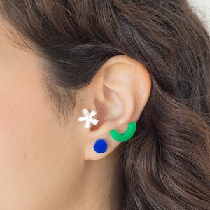 May include: A person's ear with three earrings. The top earring is a silver star, the middle earring is a blue circle, and the bottom earring is a green half-circle.