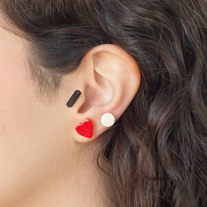 May include: A person's ear with three earrings: a black bar, a white circle, and a red heart.