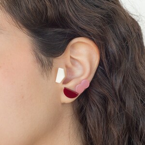 May include: A close-up of a person's ear with three geometric earrings. The earrings are a white rectangle, a pink cloud shape, and a red half-circle.