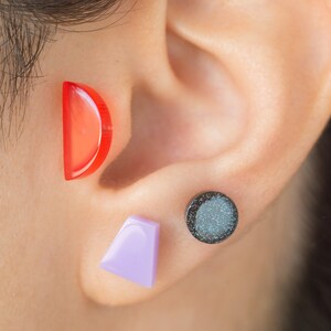 May include: A close-up of a person's ear with three different earrings. The earrings are a red half-circle, a purple triangle, and a black round earring with glitter.
