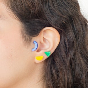 May include: A person wearing three different coloured geometric earrings. The earrings are a blue crescent moon, a yellow half circle, and a green triangle.
