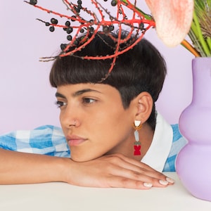 May include: A person with short, dark hair wearing a blue and white checked shirt and a pair of geometric earrings. The earrings are made of red, white and gold coloured materials. The person is looking off to the side.