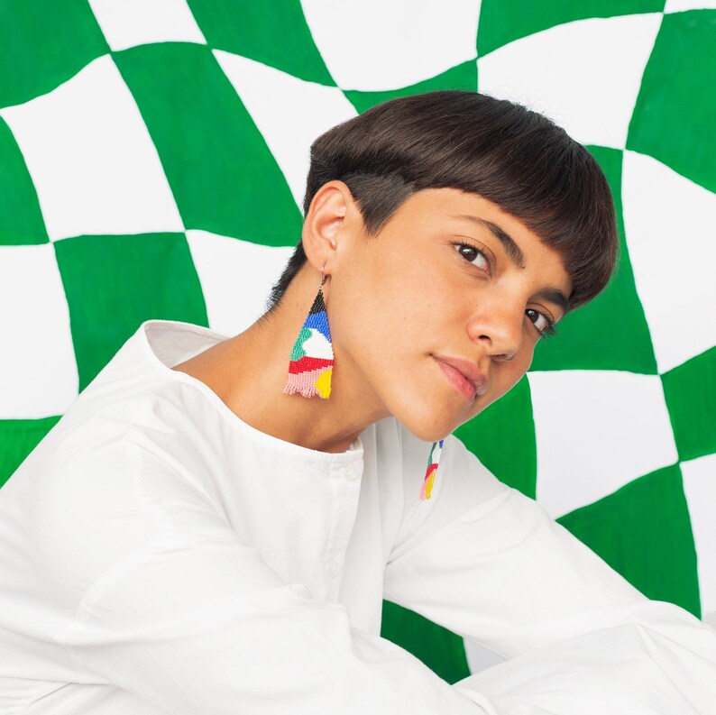 Puede incluir: Una persona con el pelo corto y casta&ntilde;o que lleva una camisa blanca y pendientes de cuentas de colores. La persona est&aacute; posando frente a un fondo a cuadros verde y blanco.