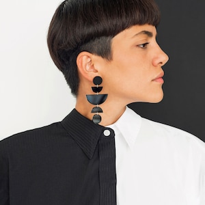 May include: Black geometric dangle earrings with gold-coloured accents. The earrings feature circles and semi-circles. The person in the image is wearing a black and white collared shirt.