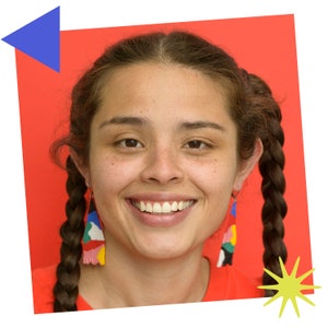 Puede incluir: Una mujer con trenzas largas y marrones sonr&iacute;e a la c&aacute;mara. Lleva una camisa roja y pendientes de cuentas de colores. El fondo es de color rojo s&oacute;lido.