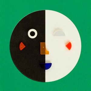 May include: A black and white circle with a face made of colorful shapes. The face has a black eye, a blue mouth, an orange nose, and two red cheeks.