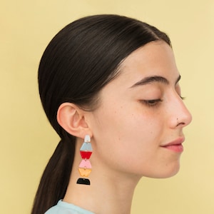 May include: A woman with long dark hair wearing a light blue shirt and colourful geometric earrings. The earrings are made of multiple layers of different coloured plastic.