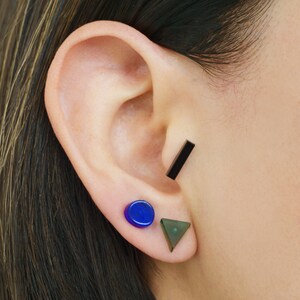 May include: A person's ear with three earrings. The top earring is a black bar, the middle earring is a blue circle, and the bottom earring is a green triangle.