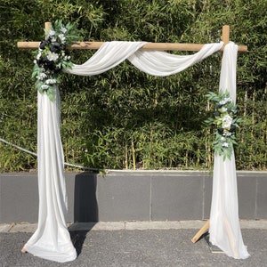 May include: White fabric draped over a wooden archway decorated with black and white flowers. The archway is set up outdoors against a backdrop of green bamboo.