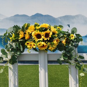 May include: A white metal archway decorated with a garland of artificial sunflowers and eucalyptus leaves. The garland is draped over the archway and hangs down on both sides.
