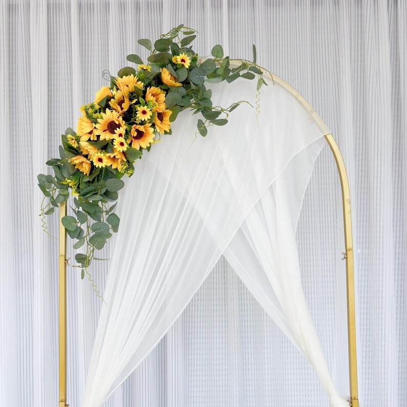 May include: A gold metal archway decorated with a garland of yellow sunflowers and green leaves. The archway is draped with a white sheer fabric.